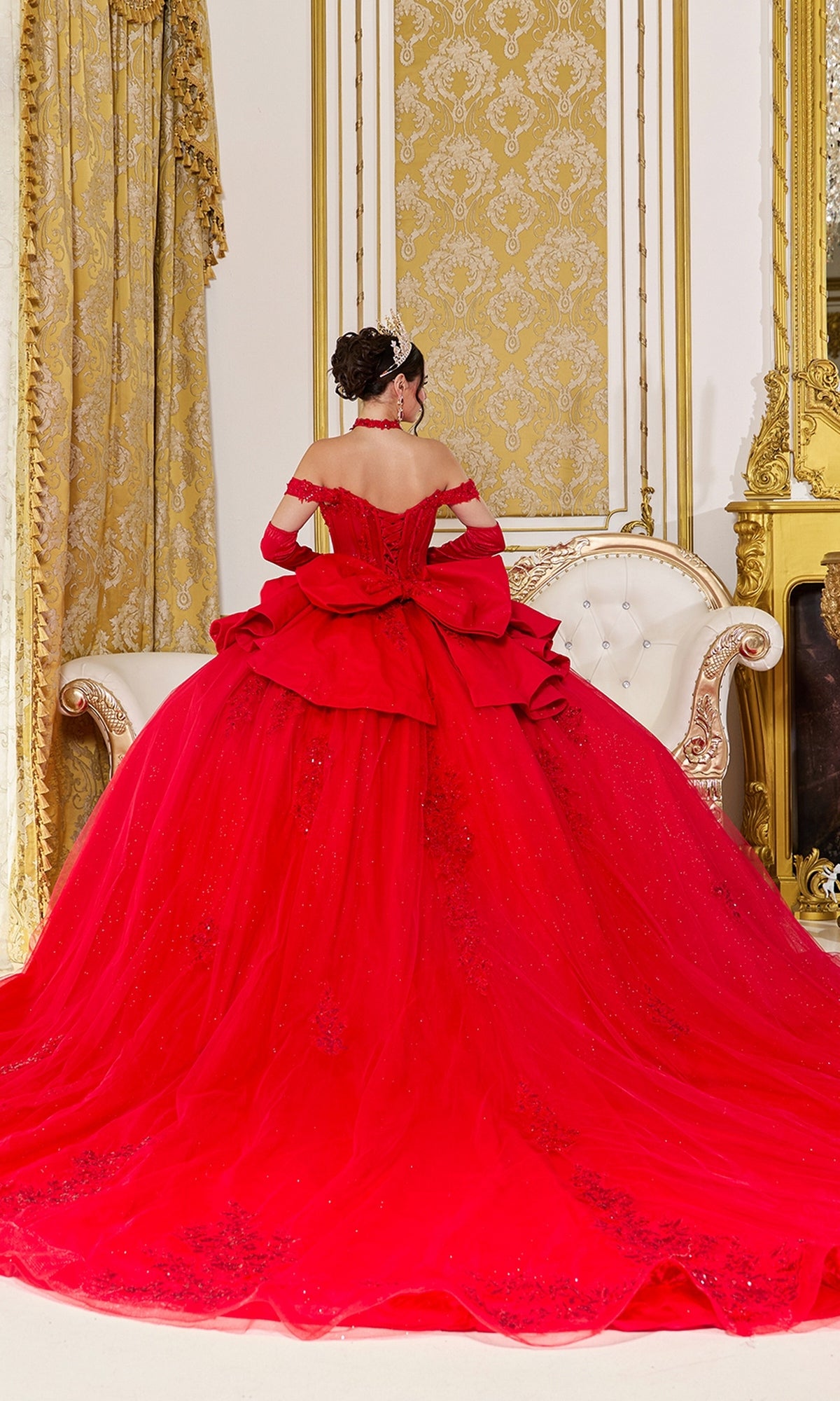 A woman wearing the Quinceanera Dress 5138 By Dancing Queen, an off-the-shoulder red ballgown, along with a tiara, sits on a white sofa seen from behind in a room adorned with ornate gold decor and patterned curtains.